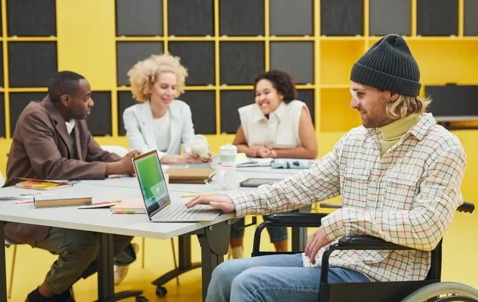 Four people are socializing around a table. Three are out of focus in the background. A Wheelchair user with a gray beanie and glasses is in focus working on his laptop.
