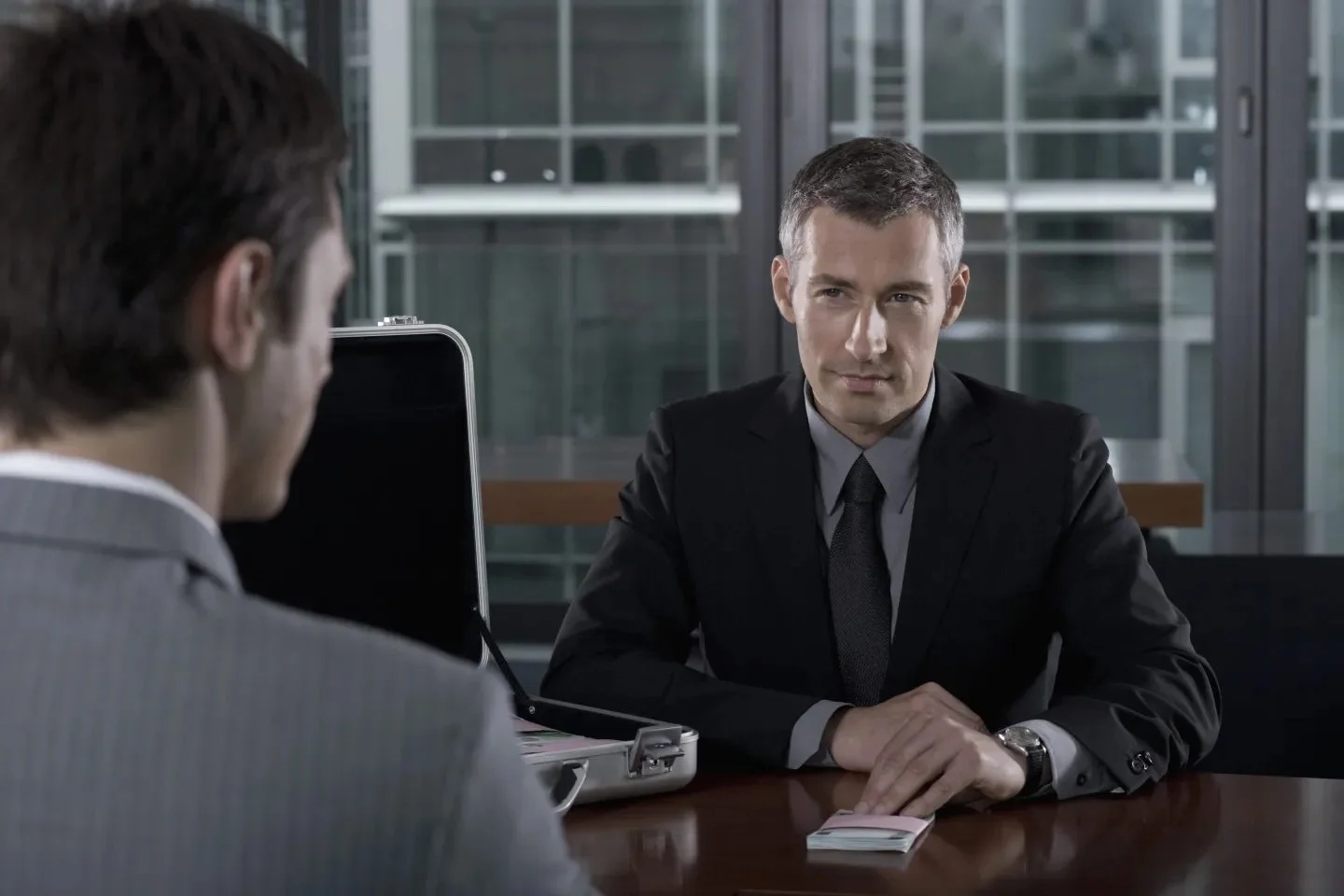 Two men in suits, sit across from each other at a conference table for a meeting.