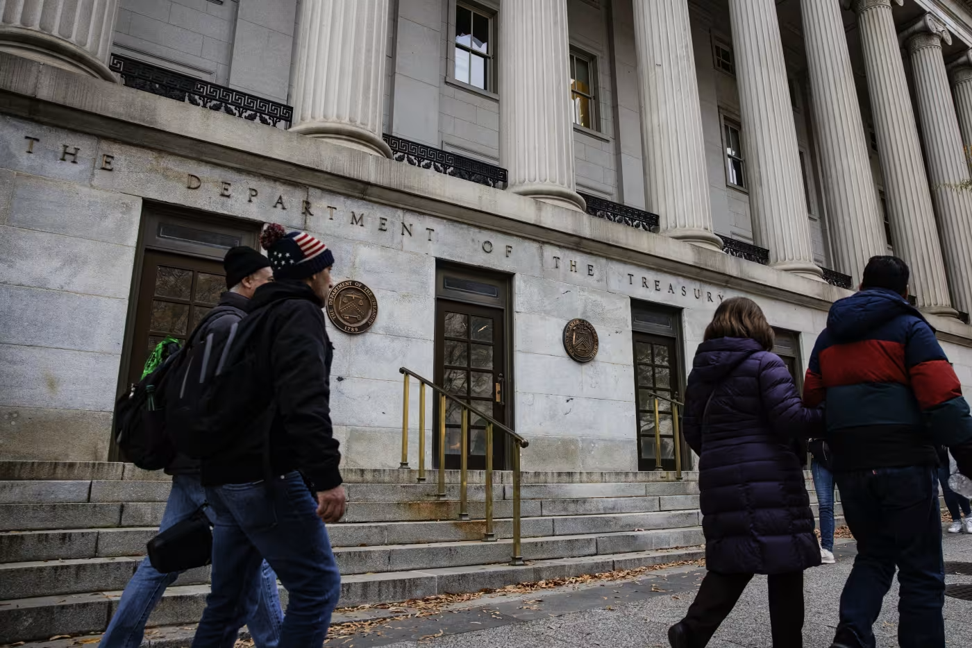People walking past the Department of the Treasury building, a historic structure with large columns and engraved signage, on a chilly day with some leaves scattered on the ground.