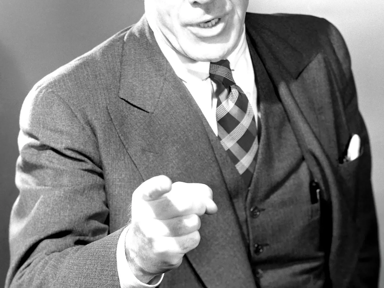 A black-and-white photo of a man in a suit pointing directly toward the camera, mid-sentence, conveying authority or confrontation.