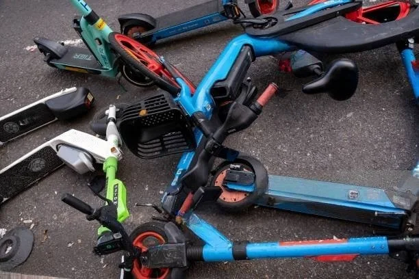 A series of E-bikes and E-scooters strewn about on the ground obstructing walkways.