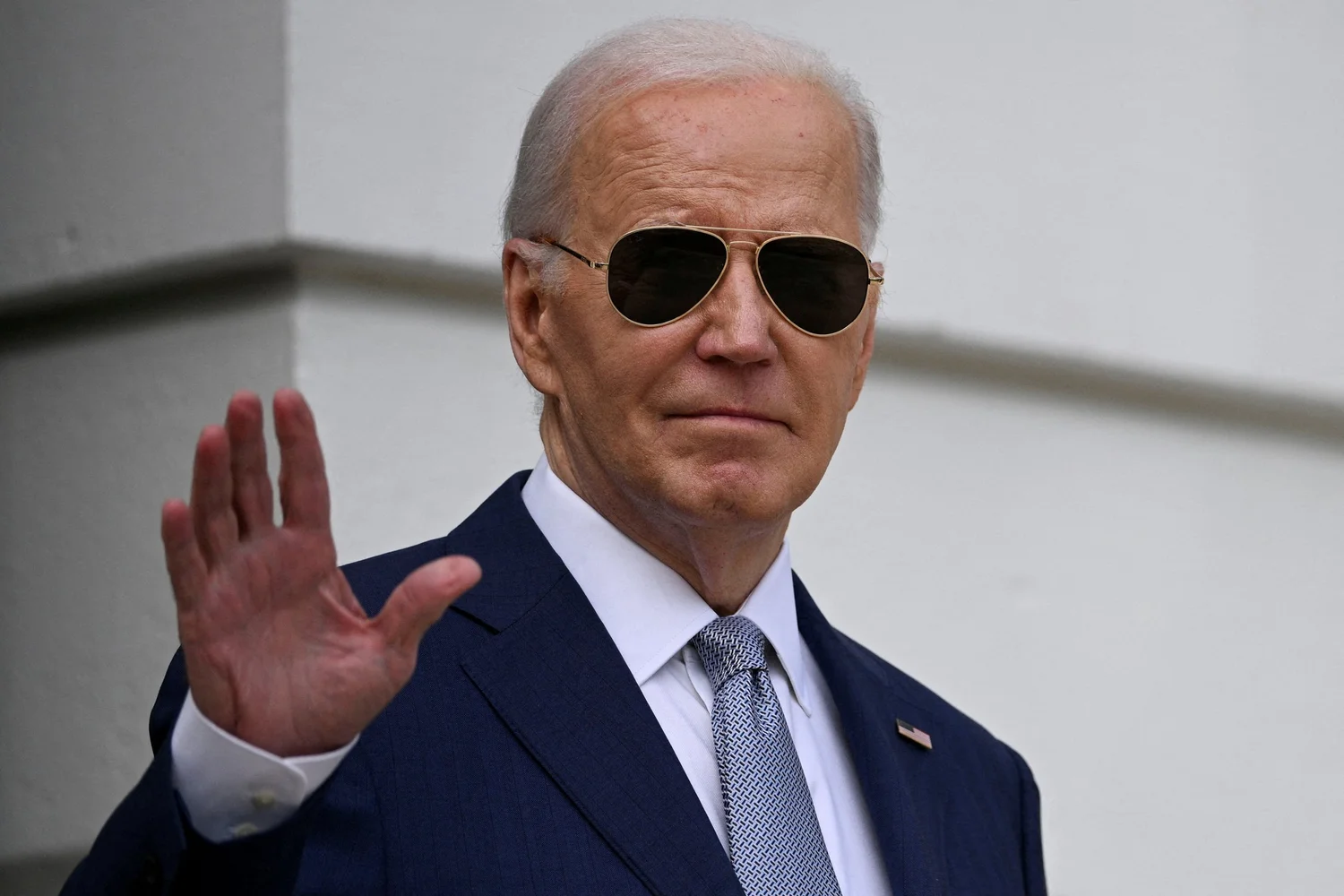 President Joe Biden, wearing a dark suit, light blue tie, and aviator sunglasses, waves to the camera.