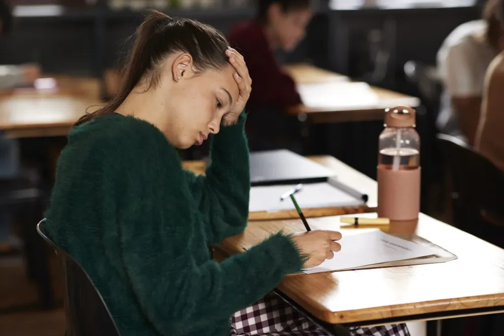 Female college student sits in a library composing a paper. She appears tired with her hand on her forehead.
