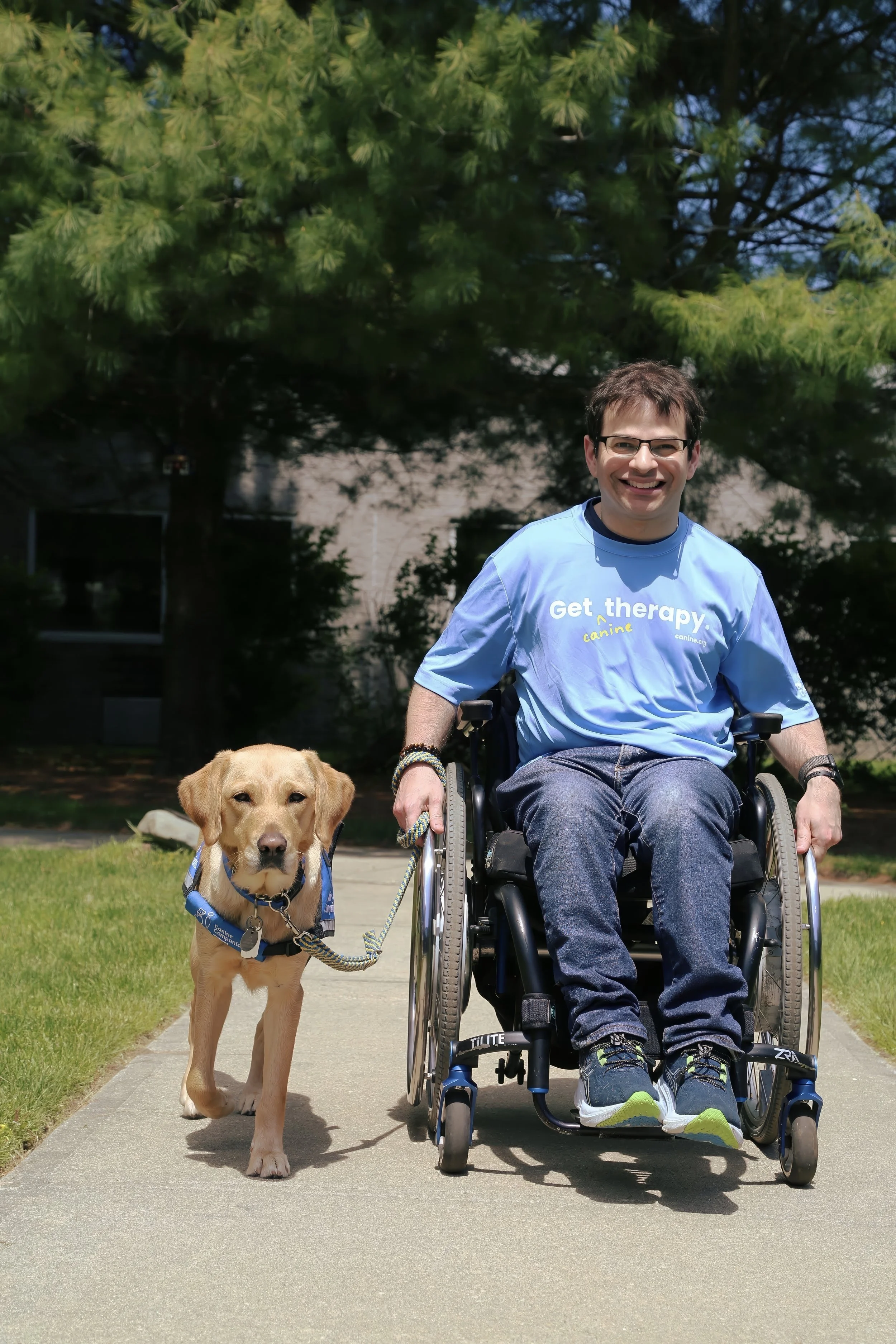 Ryan Honick smiling as he moves along an outdoor path in his wheelchair with Lovey, his yellow Labrador service dog, walking beside him