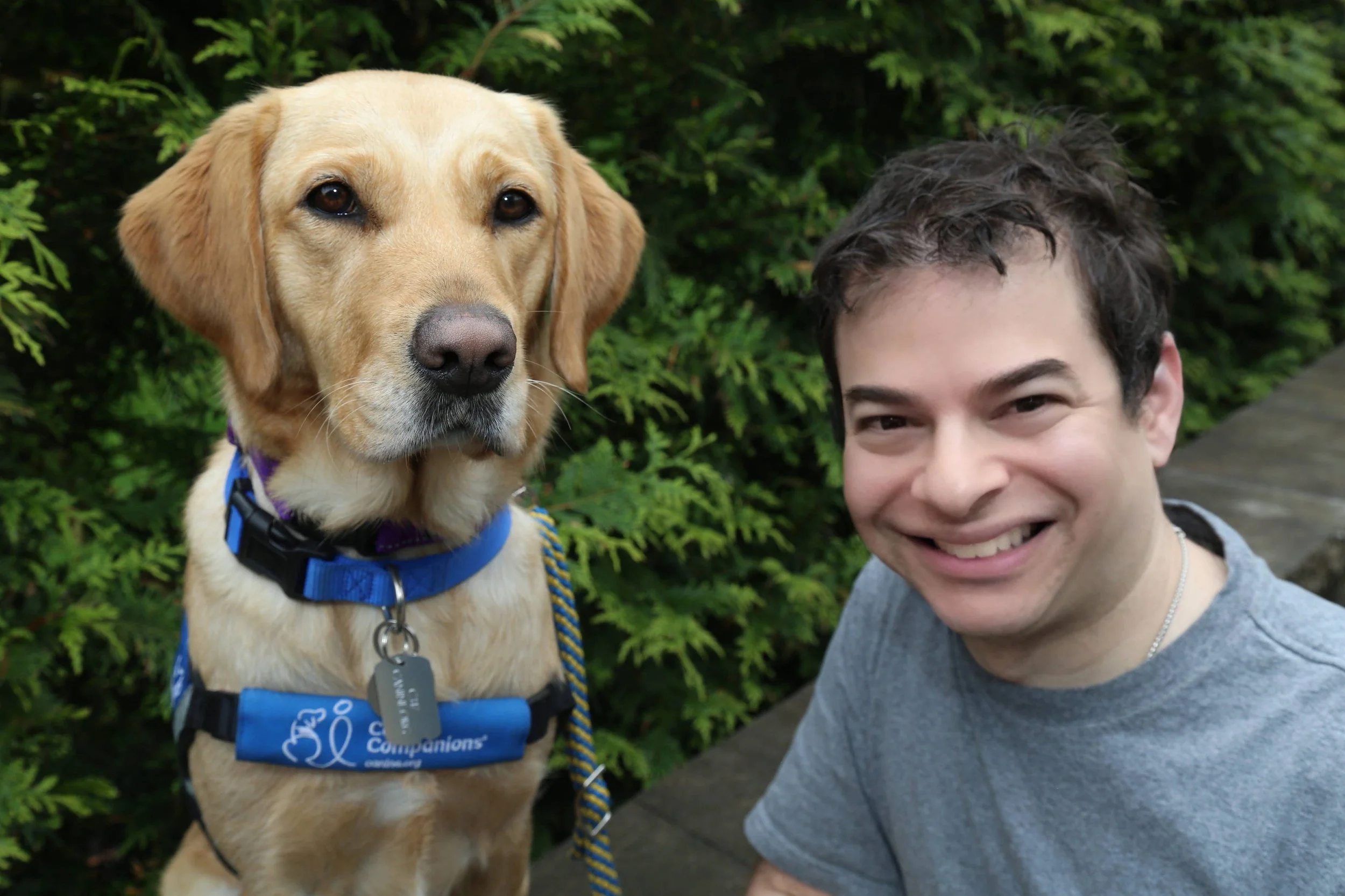 Ryan Honick smiling outdoors beside Lovey, his yellow Labrador service dog
